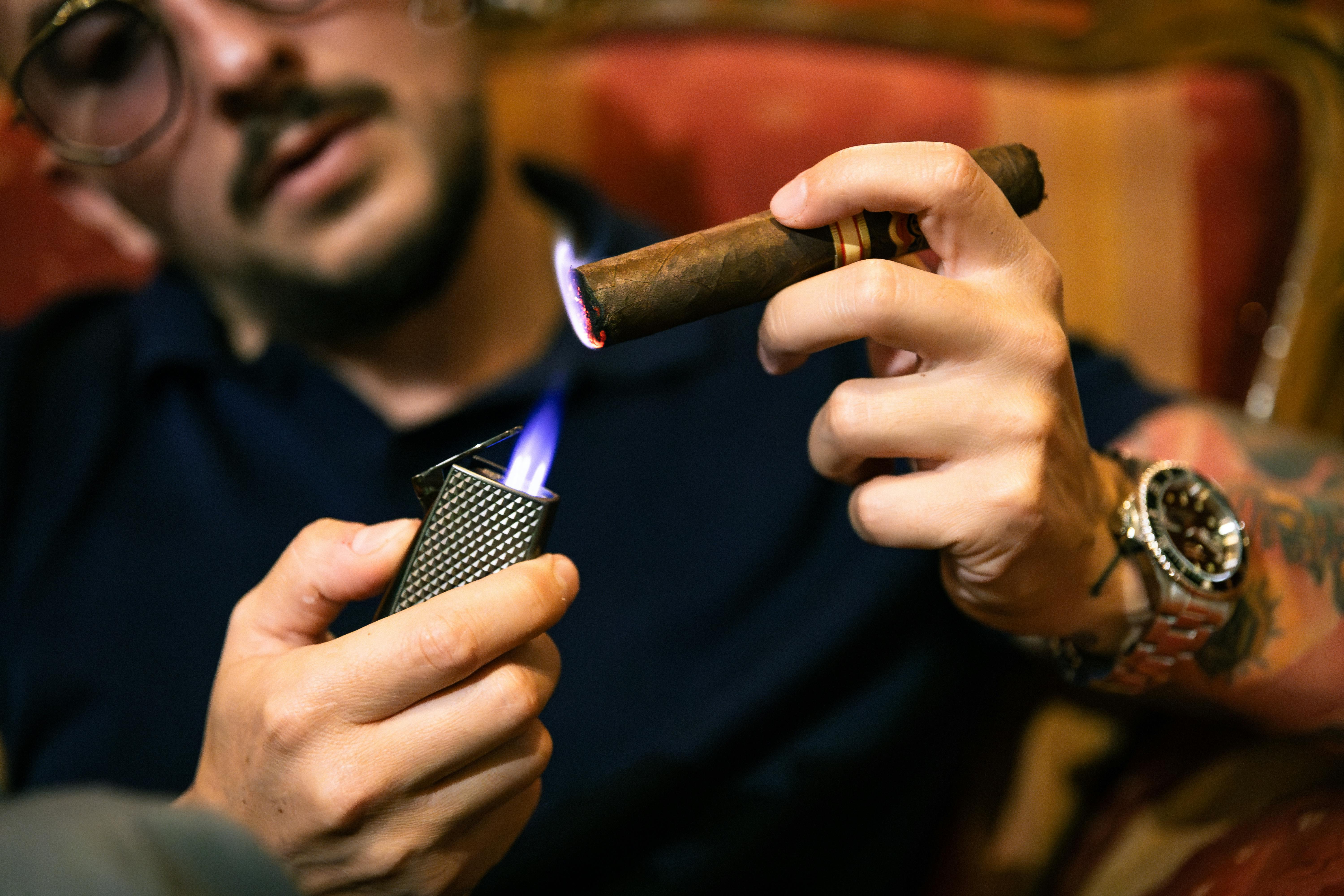 Close-up of a man lighting a cigar using a blue flame lighter, showcasing luxury and leisure.