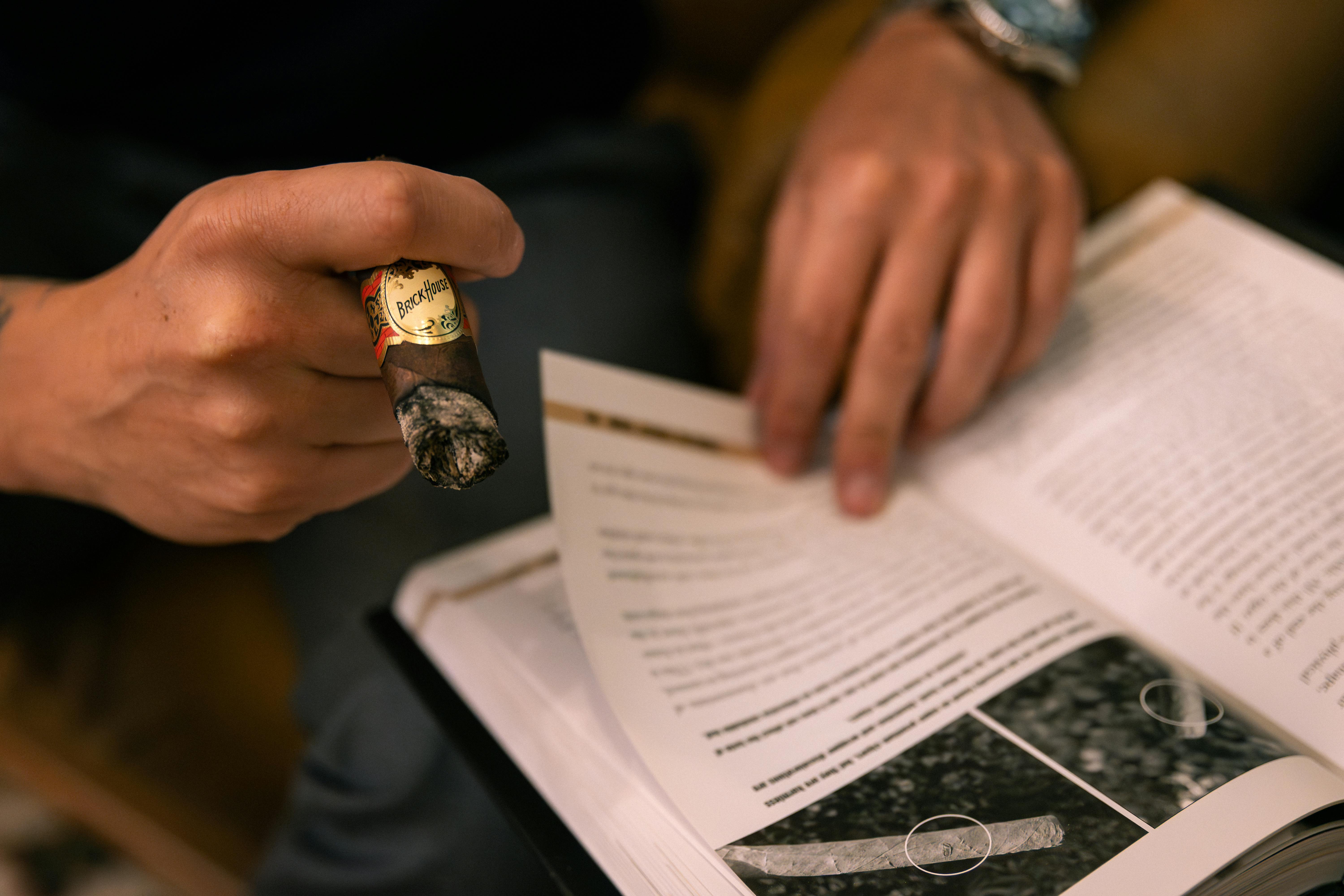 A close-up of a man's hands holding a cigar and an open book indoors.