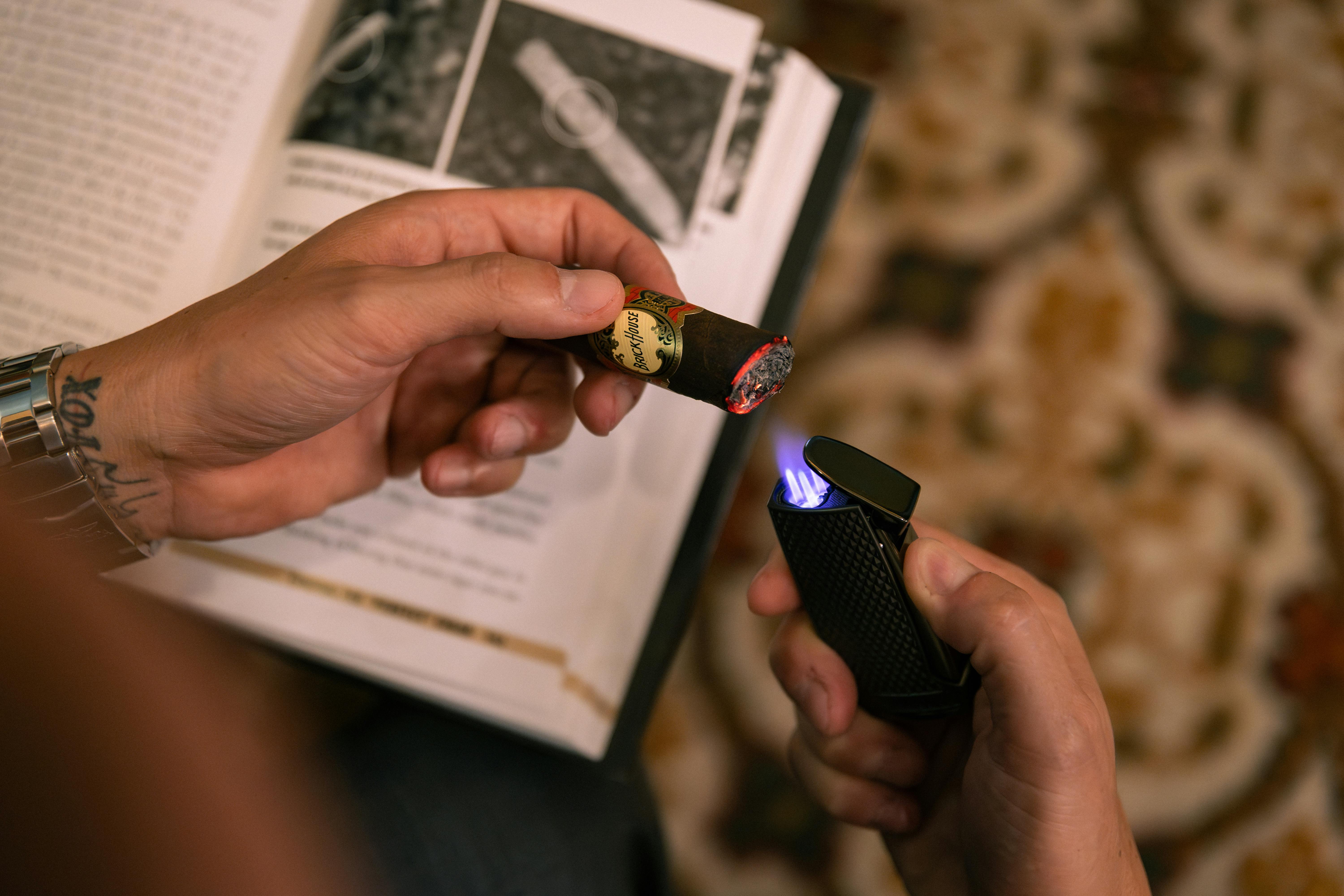 Free Close-up of hands lighting a cigar with a sleek lighter over an open book on a patterned rug. Stock Photo