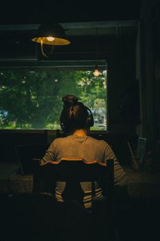 A person wearing headphones in a dim room, silhouetted against a window showing greenery outside.