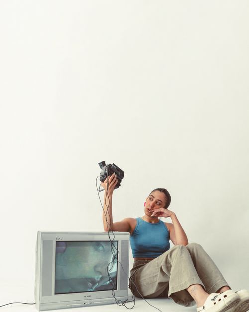 Free Woman sitting by vintage TV holding camera. Casual indoor setting with minimalist aesthetic. Stock Photo