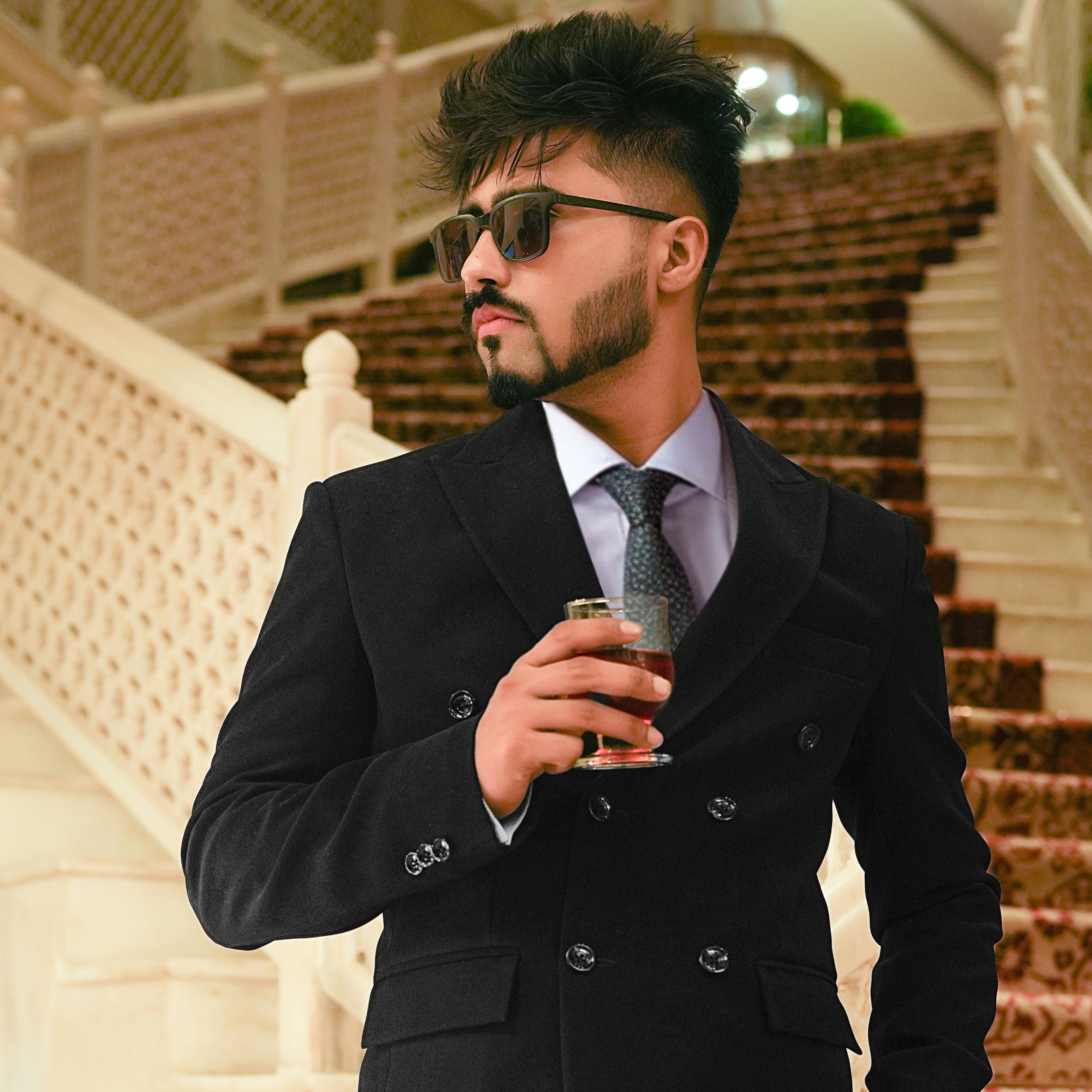 Stylish man in double-breasted suit holding a drink on a staircase, exuding confidence.