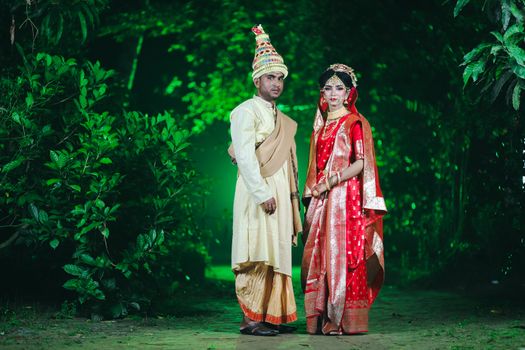 A couple in traditional Indian wedding attire poses outdoors in a lush green forest.