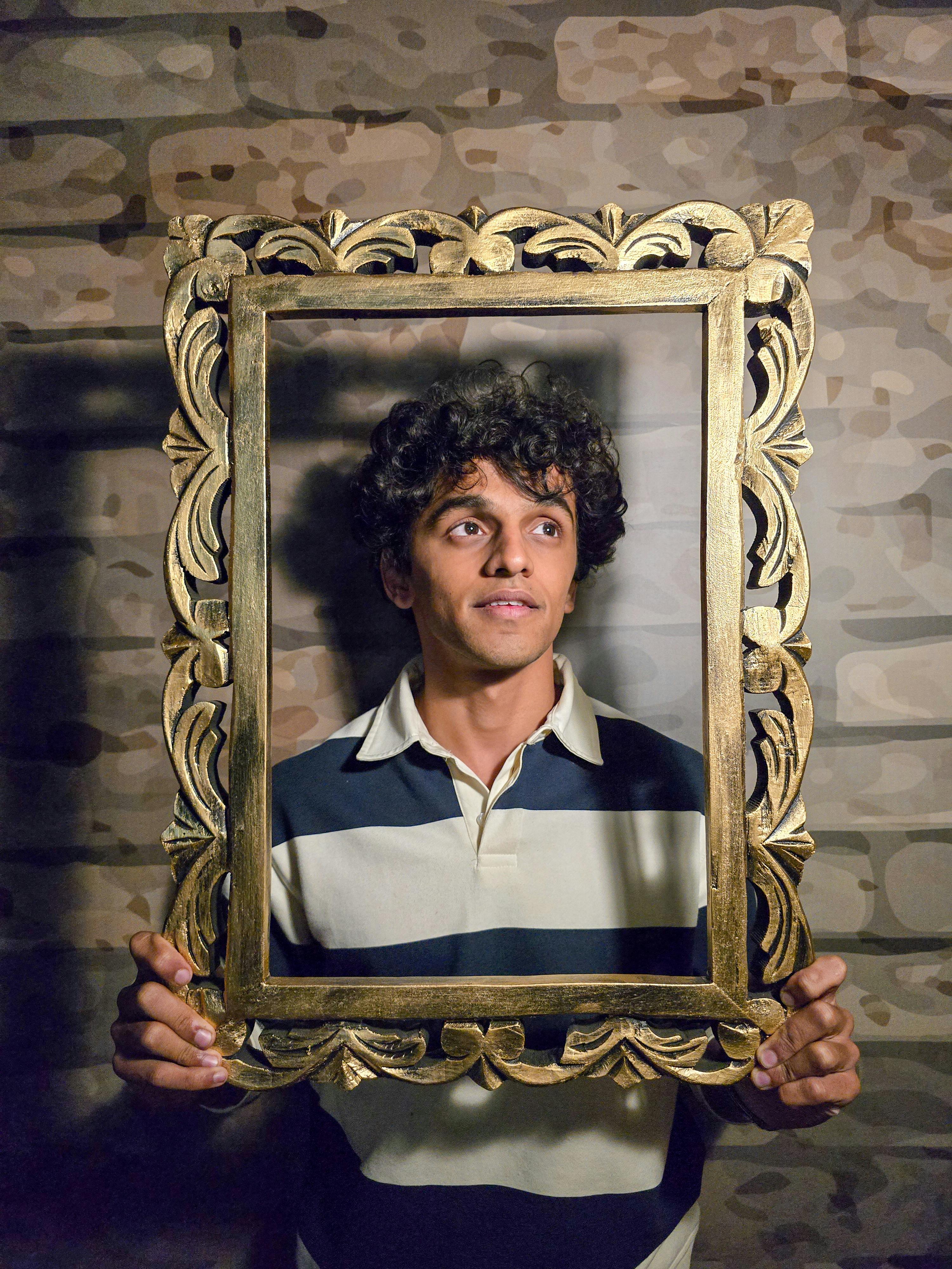 A young man with curly hair holds an ornate gold frame against a textured wall.