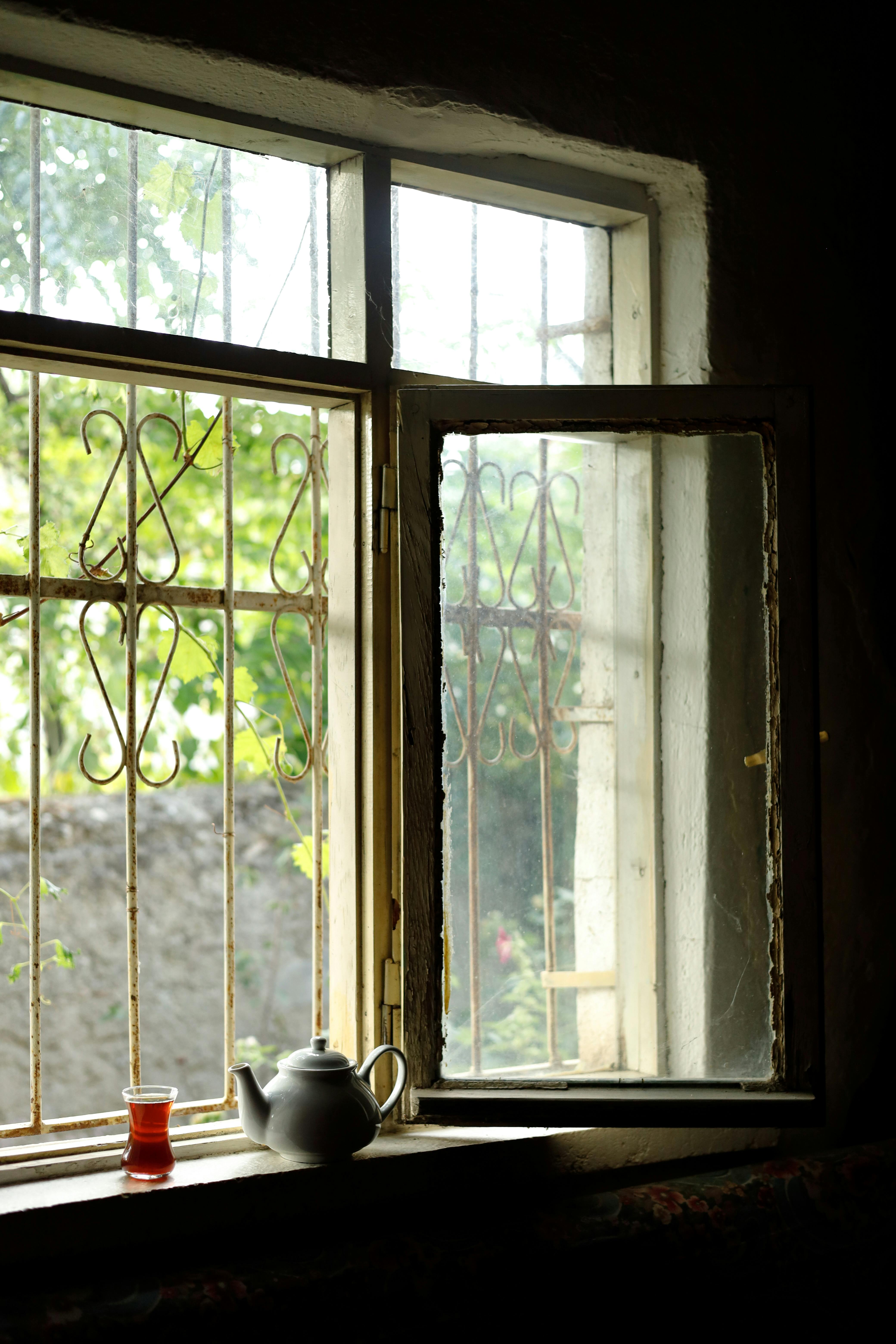 Sunlit window with teapot and glass, creating a cozy vintage atmosphere indoors.