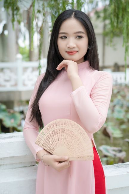 Young Vietnamese woman in traditional ao dai posing gracefully outdoors with a hand fan.