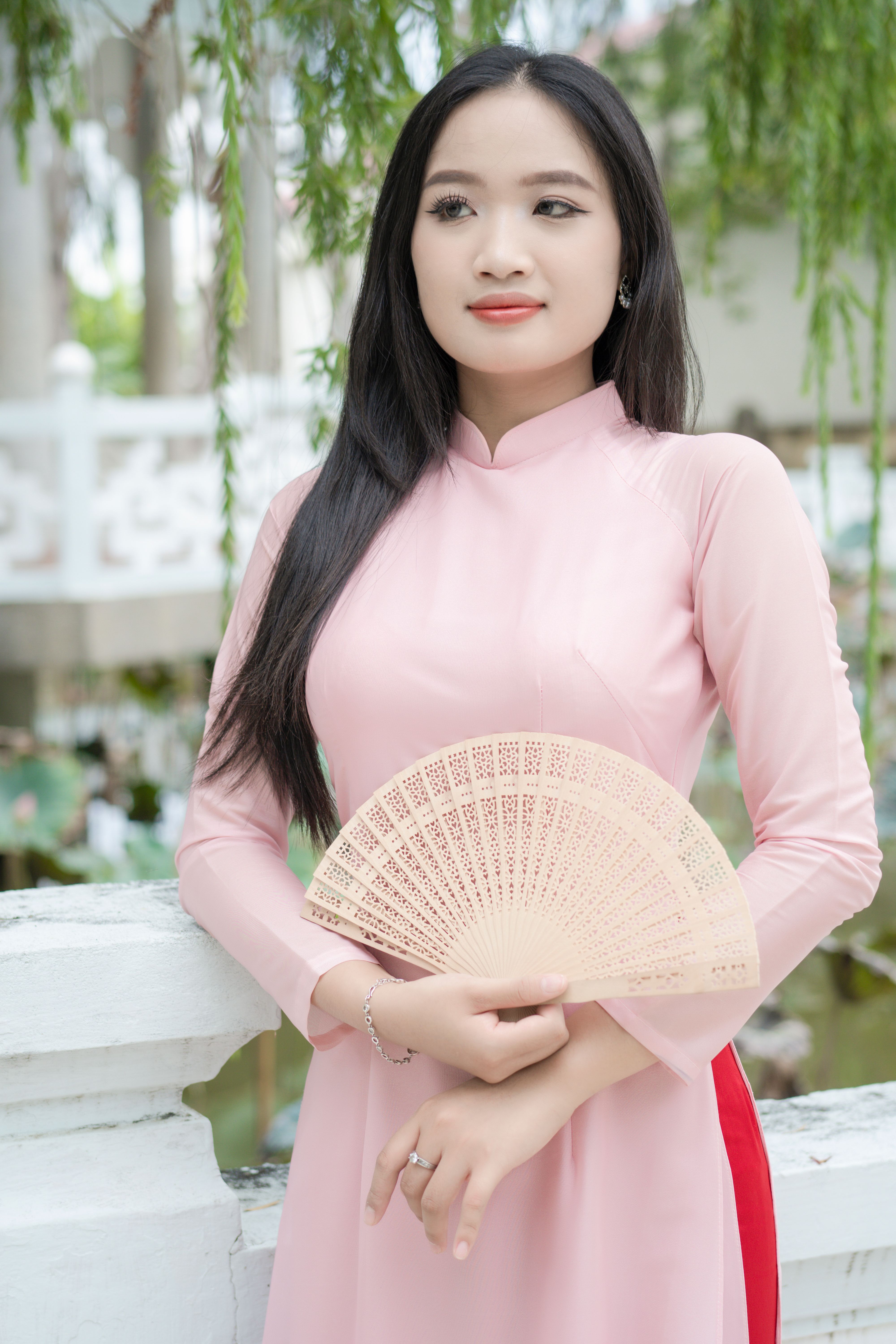 Young woman in a pink ao dai holding a fan, standing outdoors.