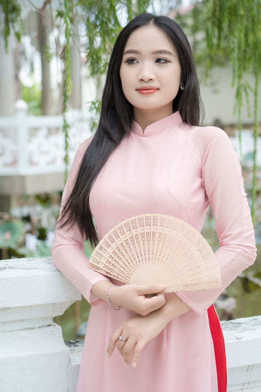 Young woman in a pink ao dai holding a fan, standing outdoors.