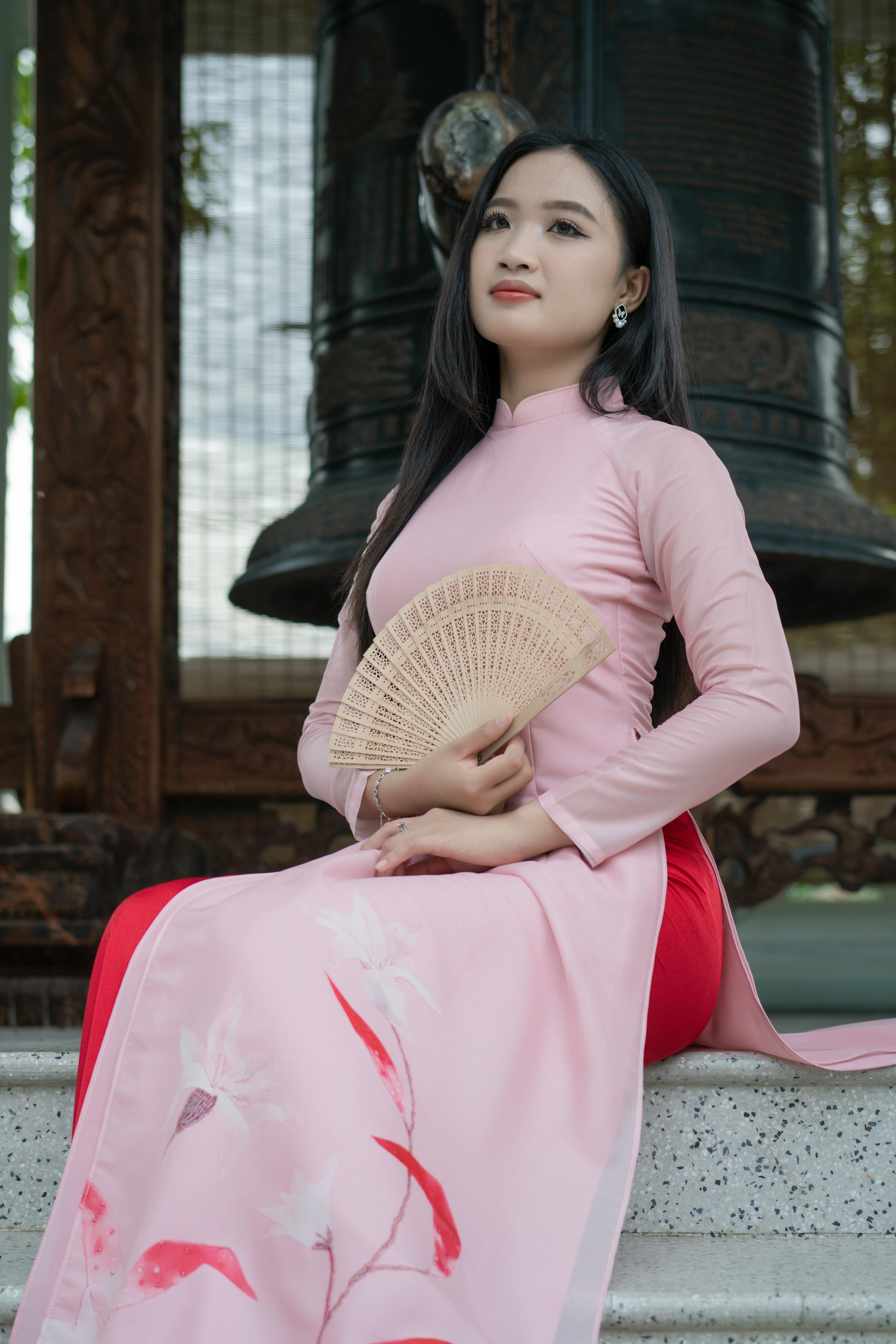 A young woman in a pink Ao Dai sits elegantly by a bell, exuding grace.
