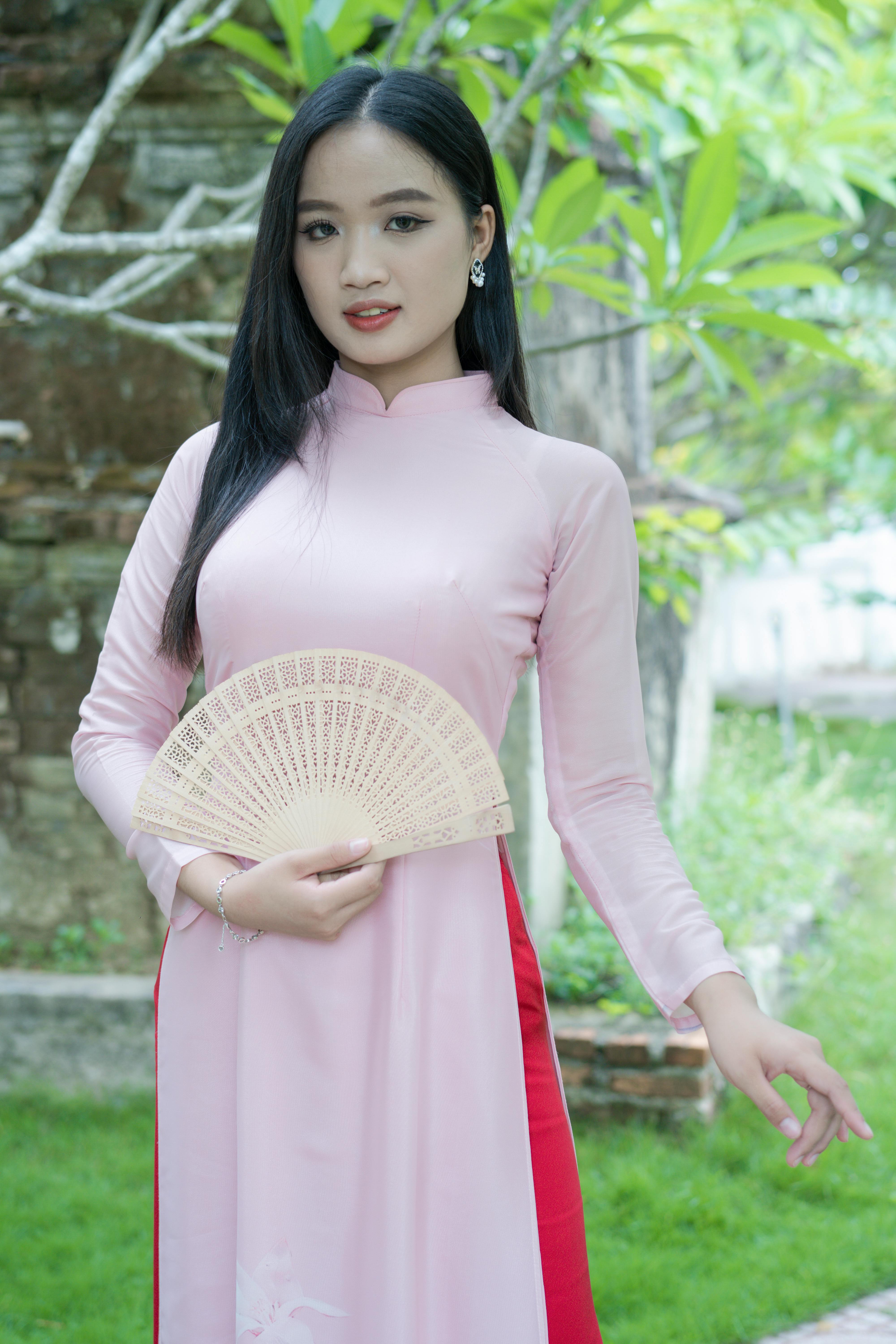 A young woman in a pink ao dai holds a fan, standing in a lush outdoor setting.
