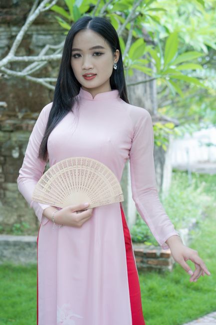 A young woman in a pink ao dai holds a fan, standing in a lush outdoor setting.