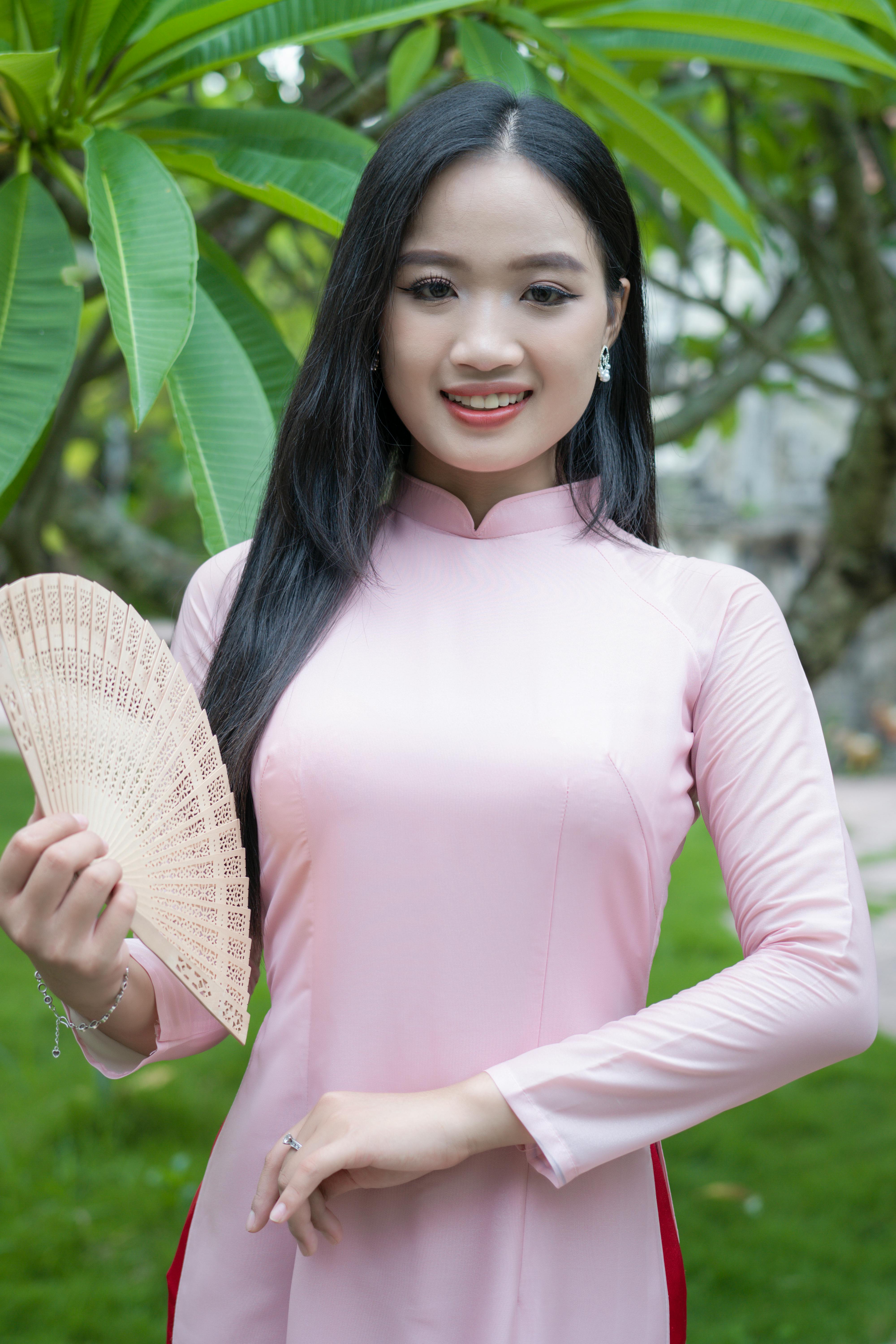 A young woman in a pink traditional dress stands outdoors holding a fan, smiling confidently.
