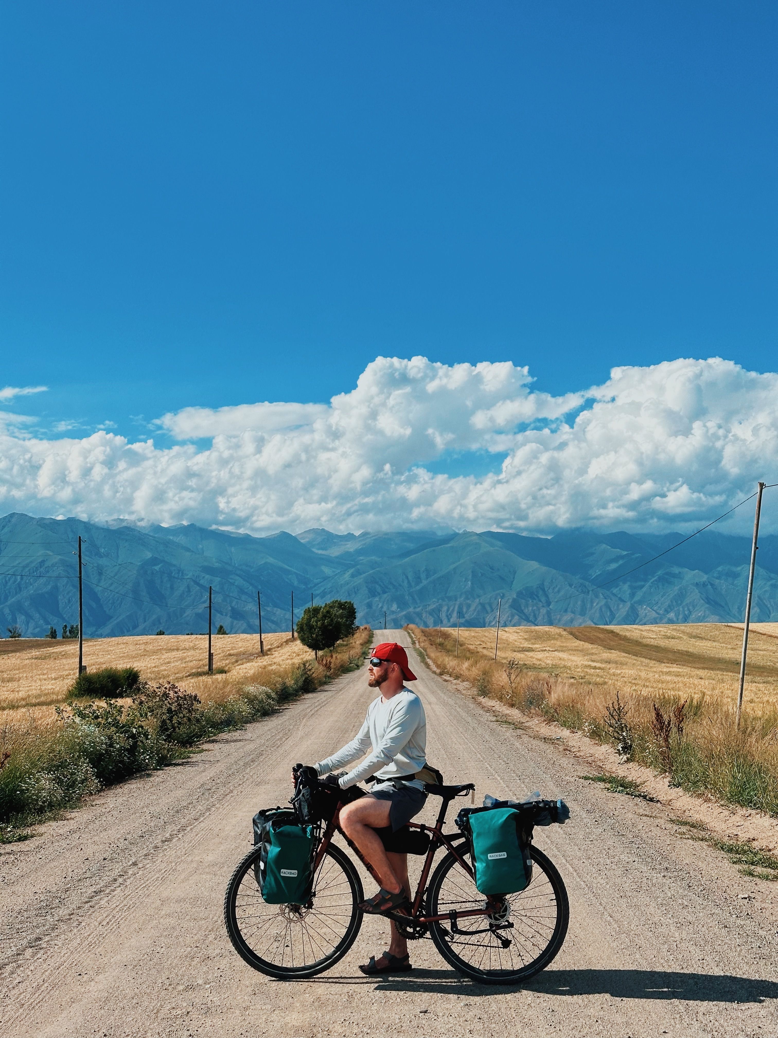 A solo cyclist rides on a scenic mountain road in Kyrgyzstan under a clear summer sky.