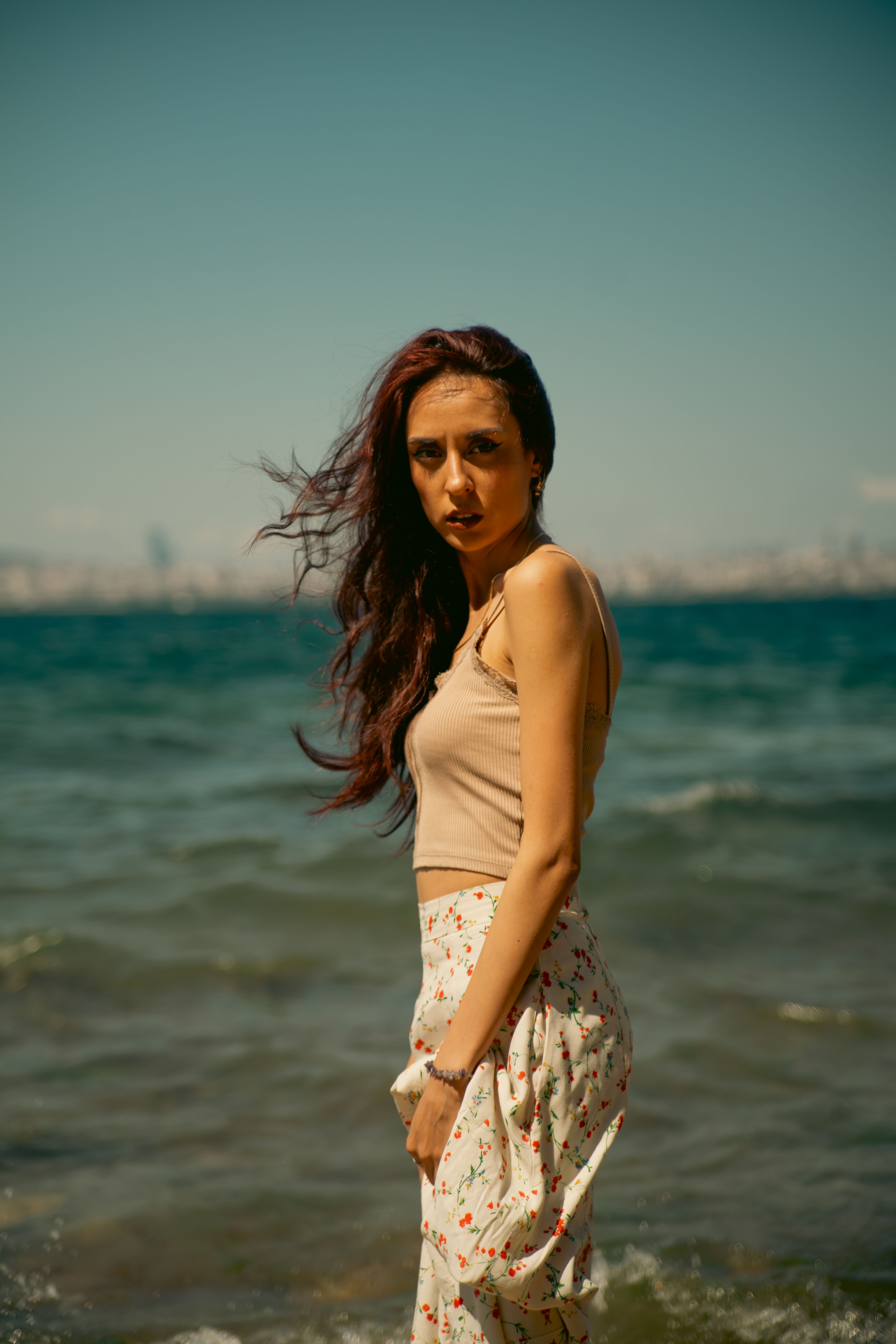 A young woman standing by the sea, with wind-swept hair under a sunny sky.