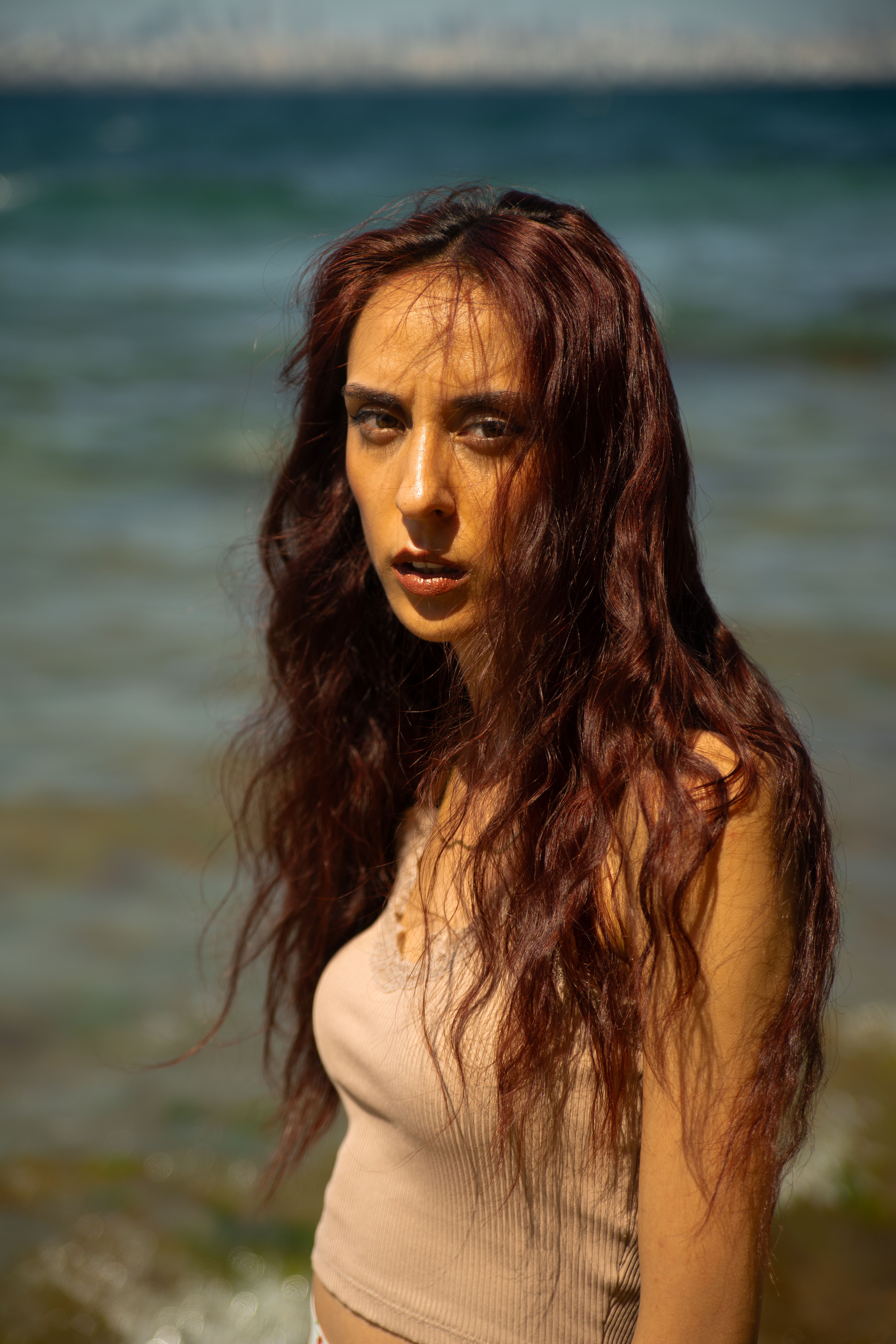 A woman with long hair stands by the sea under the sun, exuding summer vibes and natural beauty.