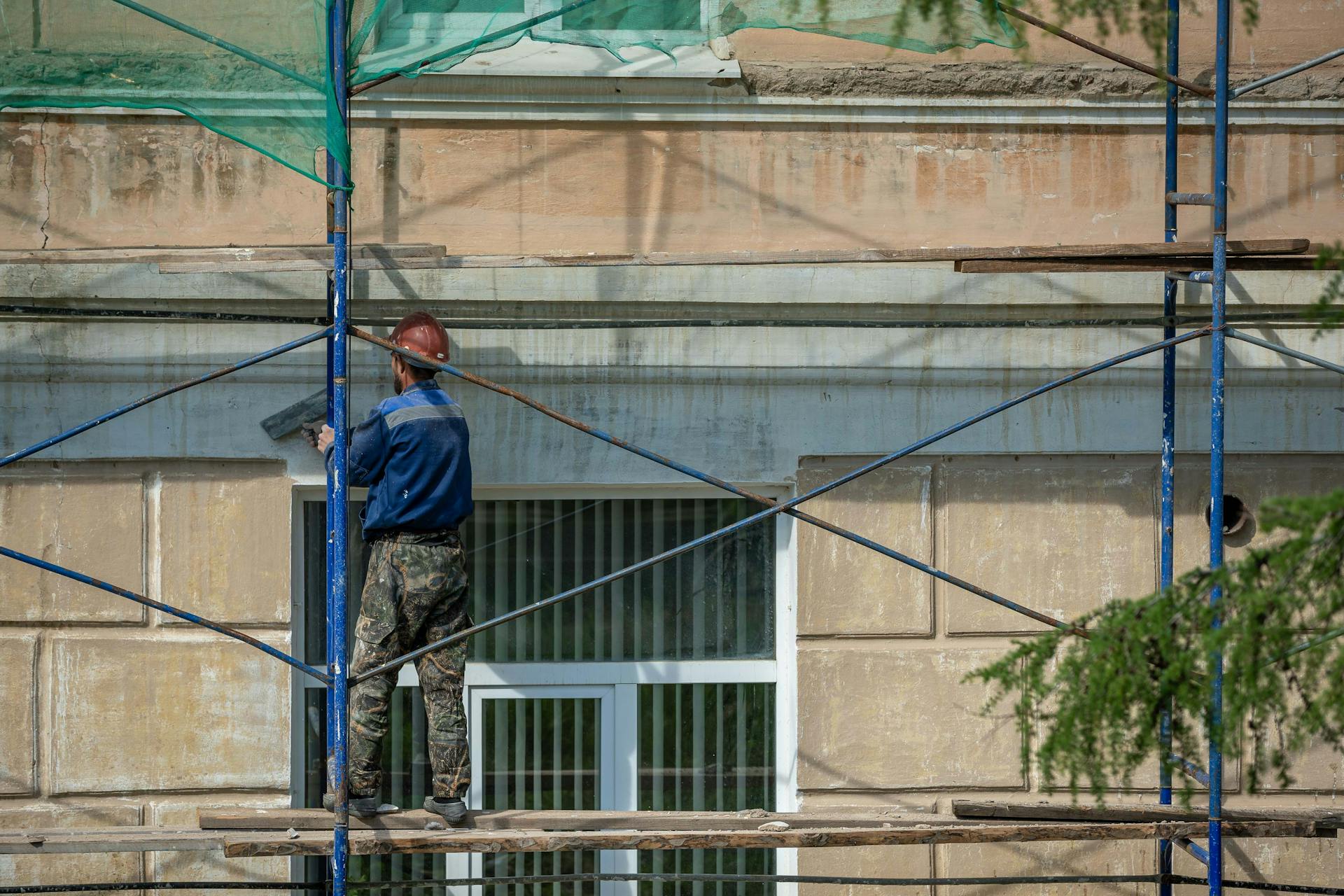 A construction worker in a hard hat repairs a building facade on scaffolding in an urban setting.