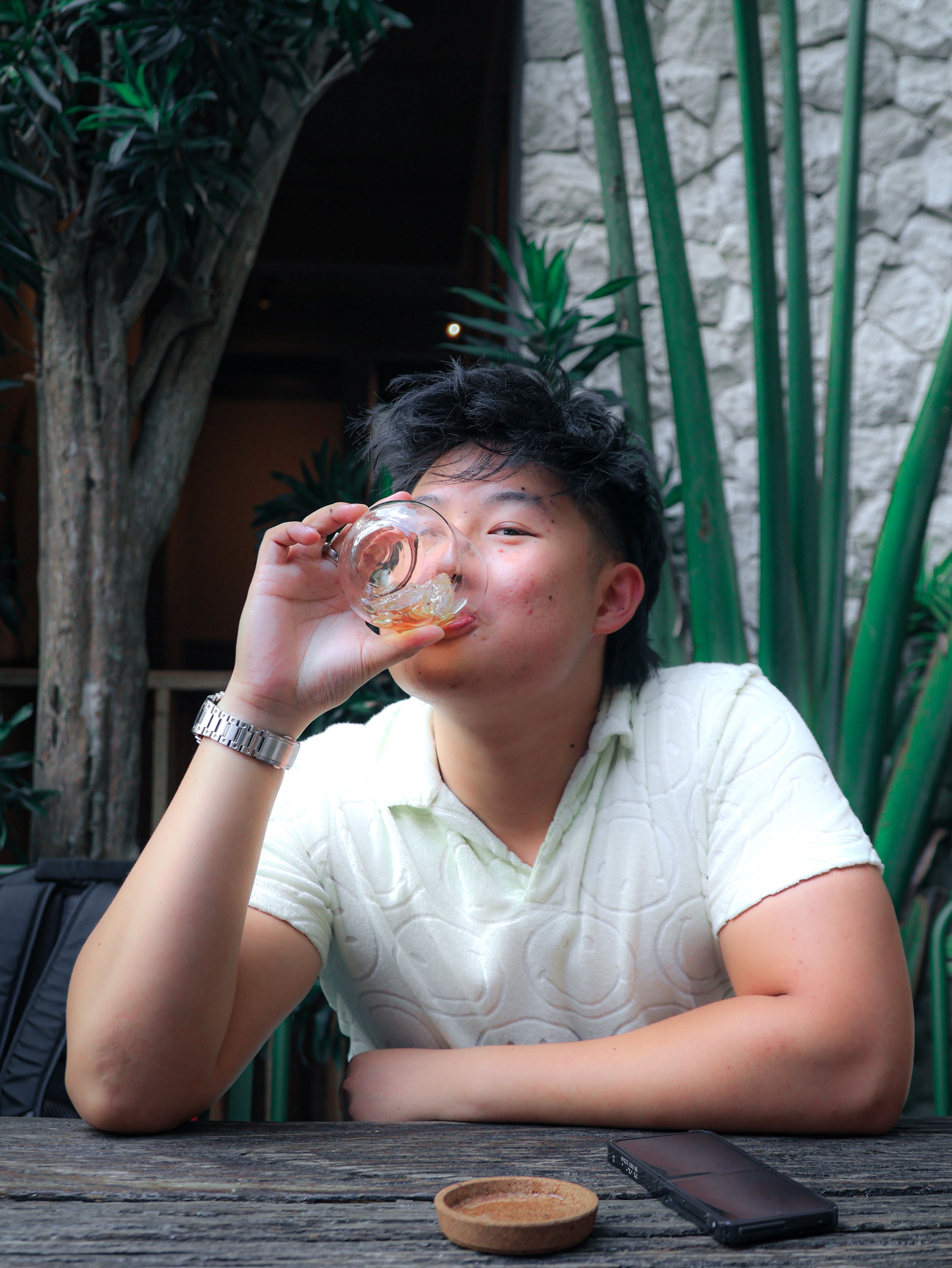 A young man savoring a drink at a rustic outdoor setting with lush greenery.