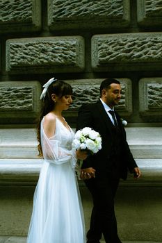A bride and groom in elegant attire walk by a historic stone wall holding hands.
