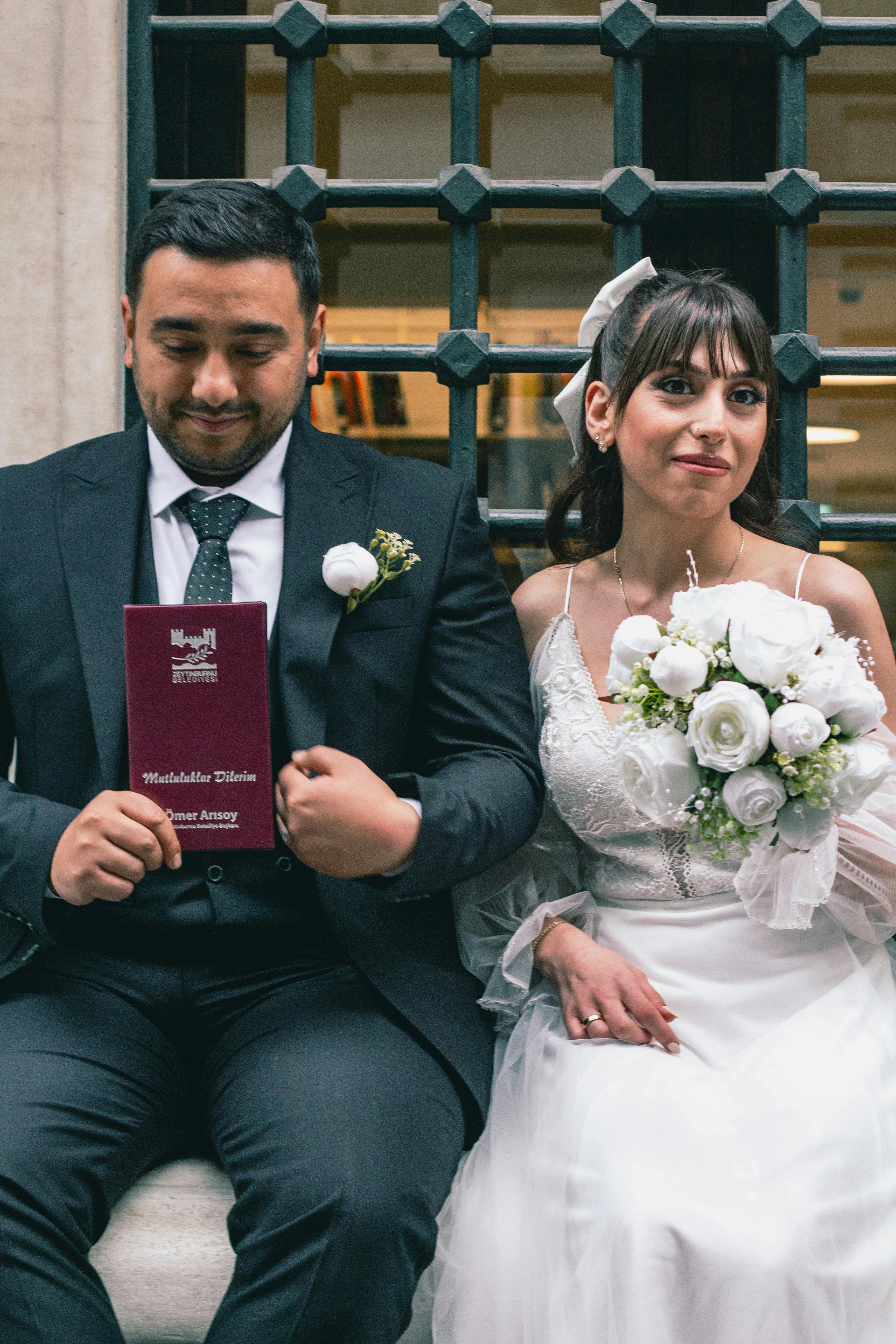 Bride and groom sitting outside a building, holding wedding bouquet and certificate.