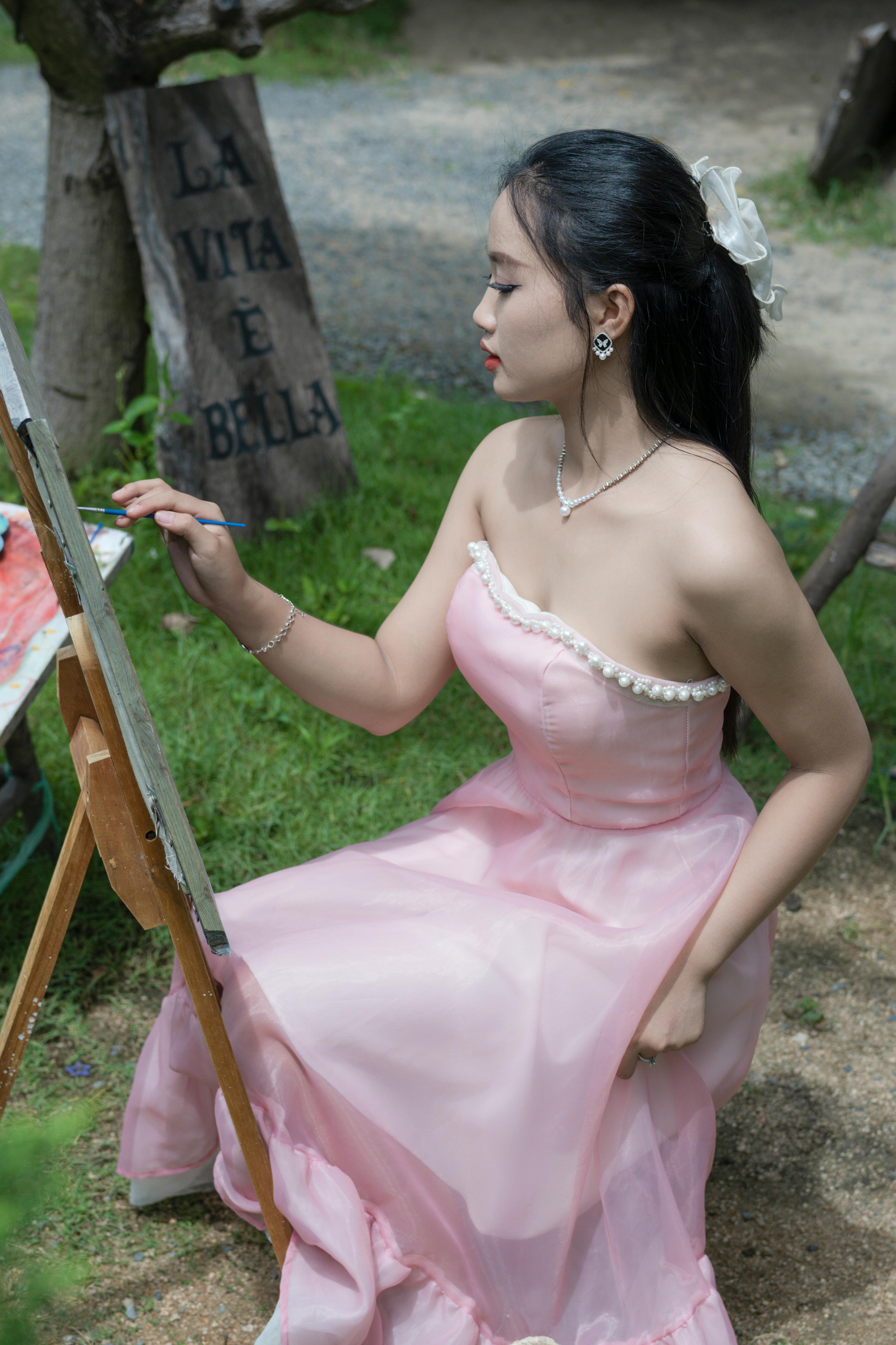 A young woman in a pink dress painting outdoors, capturing a moment of relaxation and creativity.
