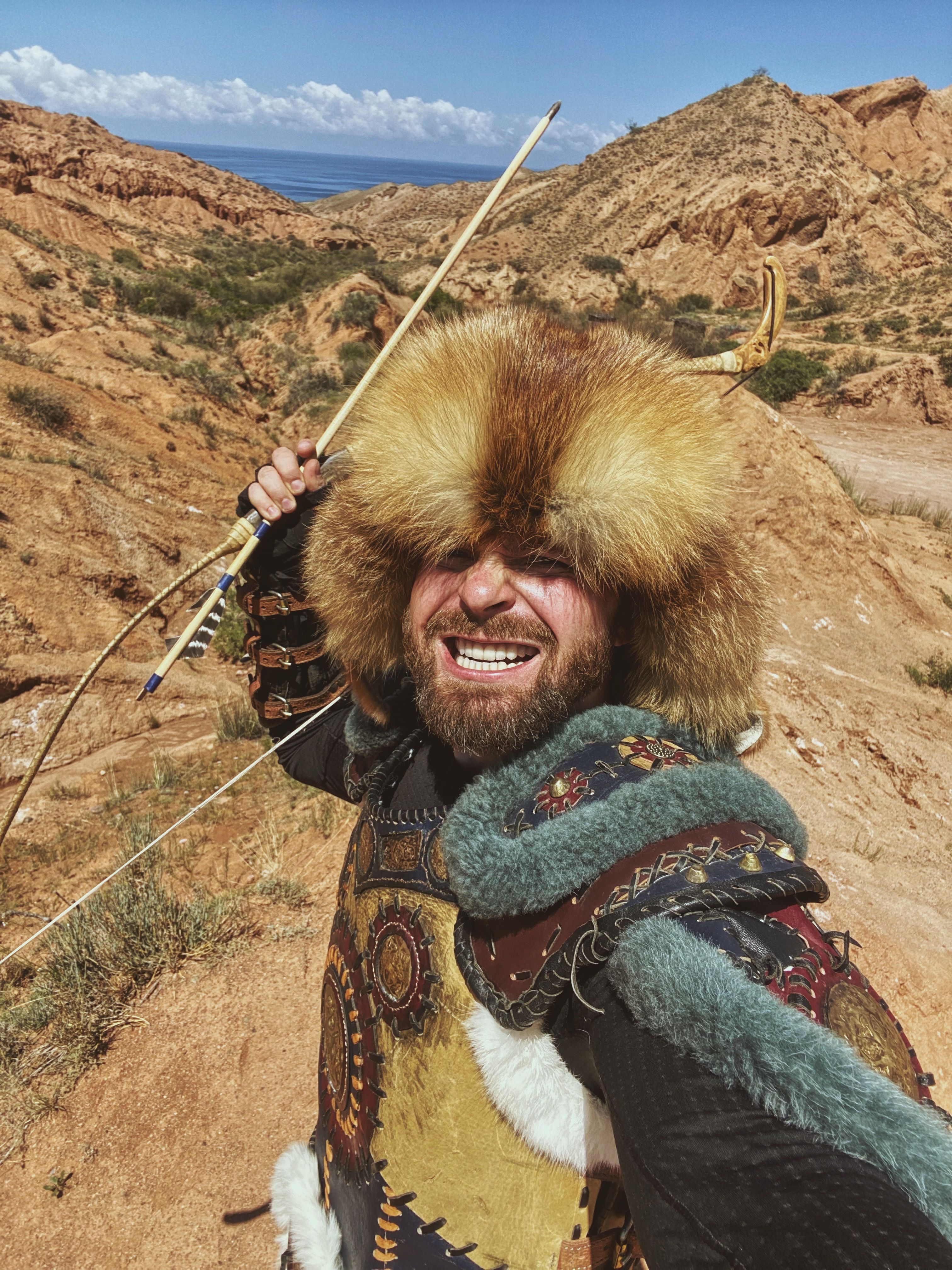 A smiling nomadic archer in traditional fur attire takes a selfie in a rugged desert landscape.