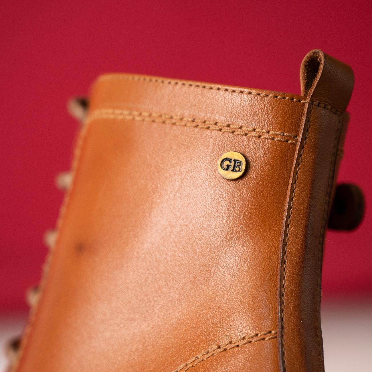 Close-up of a tan leather boot with GB logo against a red backdrop.