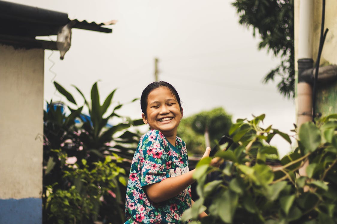 免費 閉著眼睛站在綠葉植物旁邊的小女孩微笑著的照片 圖庫相片