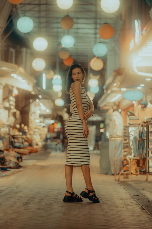 Free Young woman in striped dress poses stylishly in a vibrant, lantern-lit urban market. Stock Photo