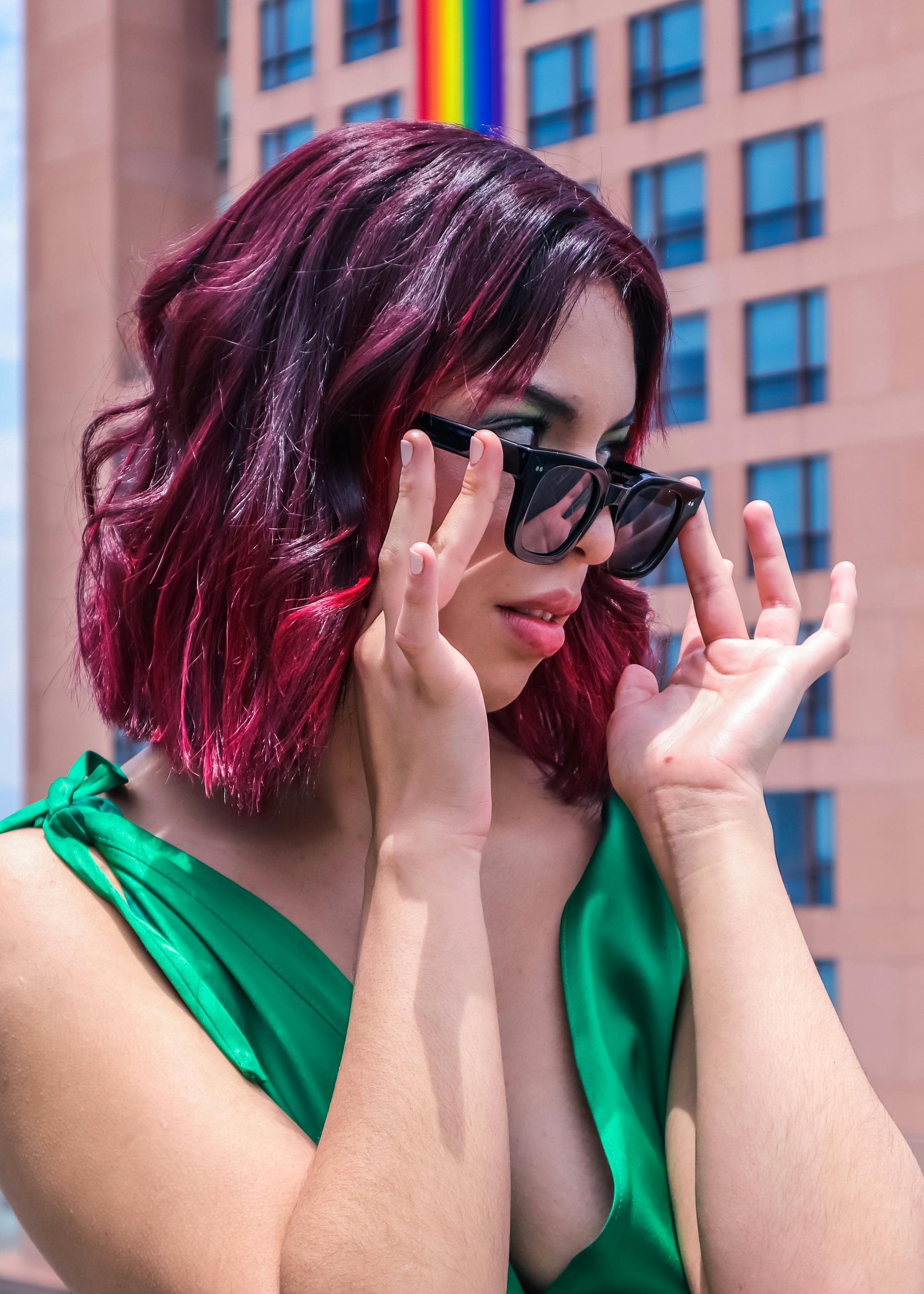 Free Young woman in a green dress adjusting sunglasses with a colorful city backdrop. Stock Photo
