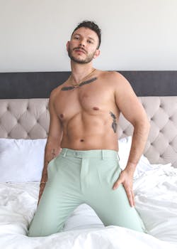 A topless man confidently poses indoors, showcasing fashion and elegance on a bed.