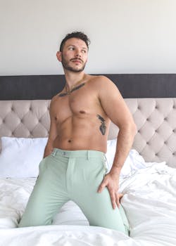 A shirtless man with tattoos pants poses confidently on a bed in a bright room.
