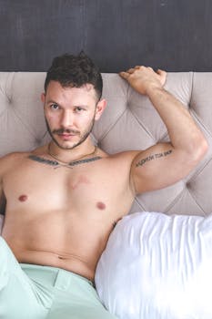 Shirtless man with tattoos relaxing on a tufted bed, conveying comfort and leisure.