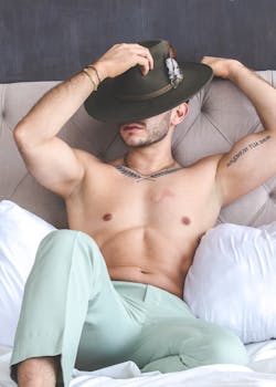 Relaxed man in green pants and hat lounging on bed, conveying elegance.