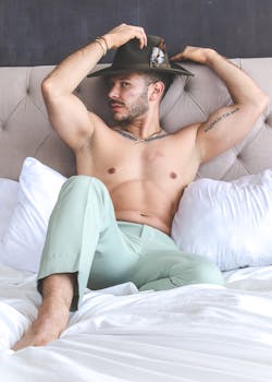 A fashionable shirtless man in green trousers and a hat reclining on a bed.