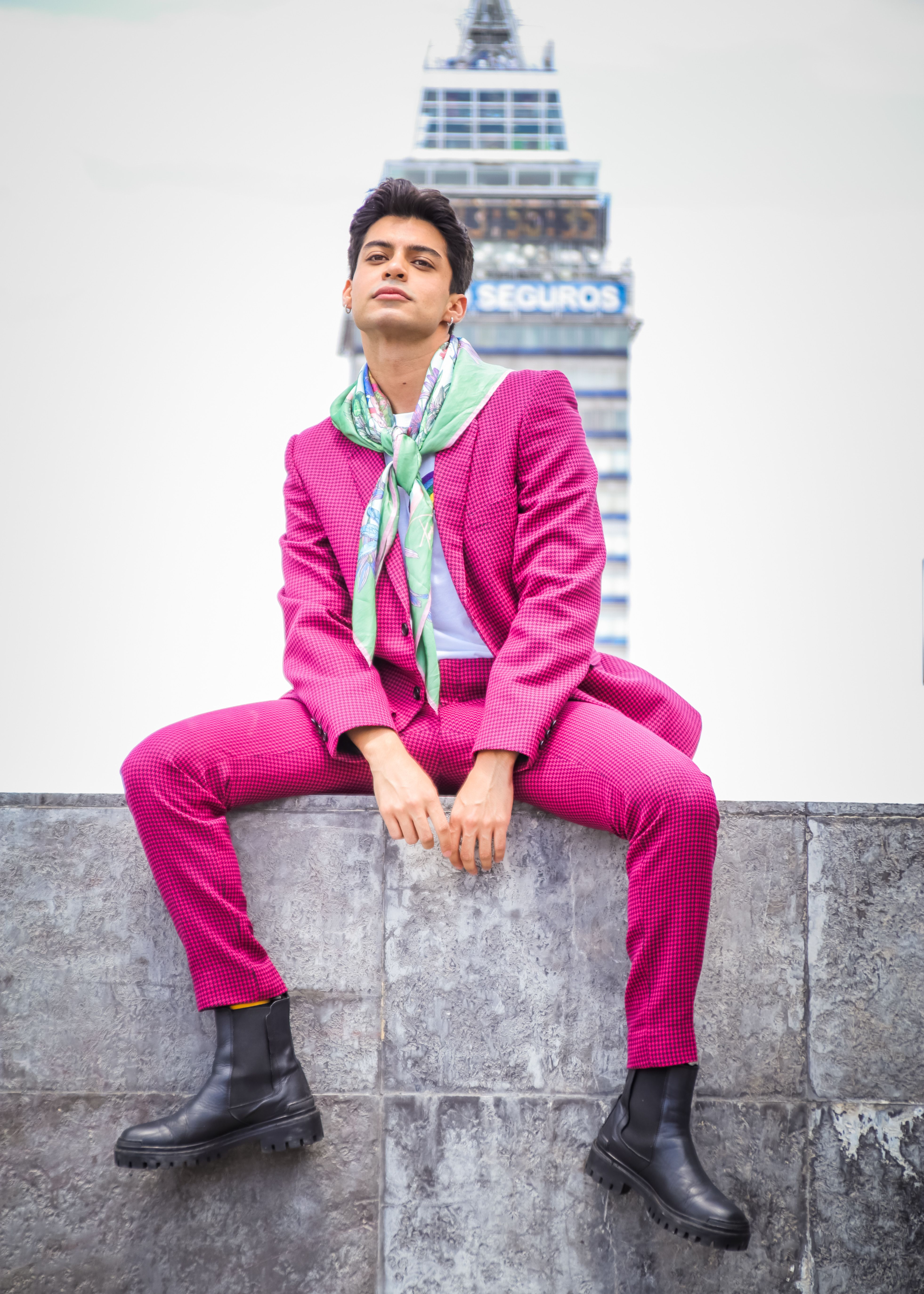 Fashionable man in a vibrant pink suit sitting with a landmark in the background.