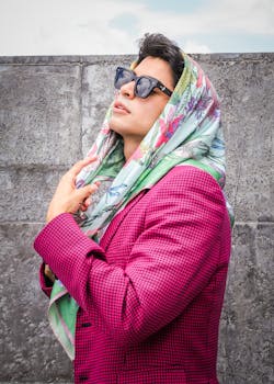 Man in pink jacket with sunglasses and scarf posing against concrete wall on a sunny day.