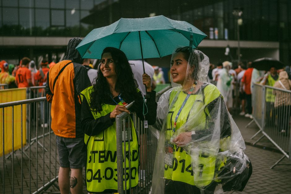 Two female stewards smiling under an umbrella at an outdoor event in rainy weather