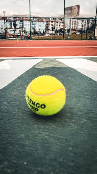 Bright yellow tennis ball on a court with urban backdrop, embodying sports and leisure.