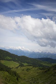 Scenic alpine landscape with rolling hills, lush greenery, and distant snowy peaks under a wide blue sky.