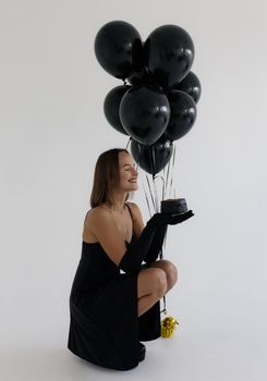 Woman in black dress holding black balloons and a small cake, symbolizing celebration and elegance.