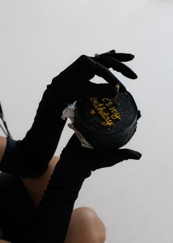 A close-up of an elegant black birthday cake held by a person wearing black gloves.