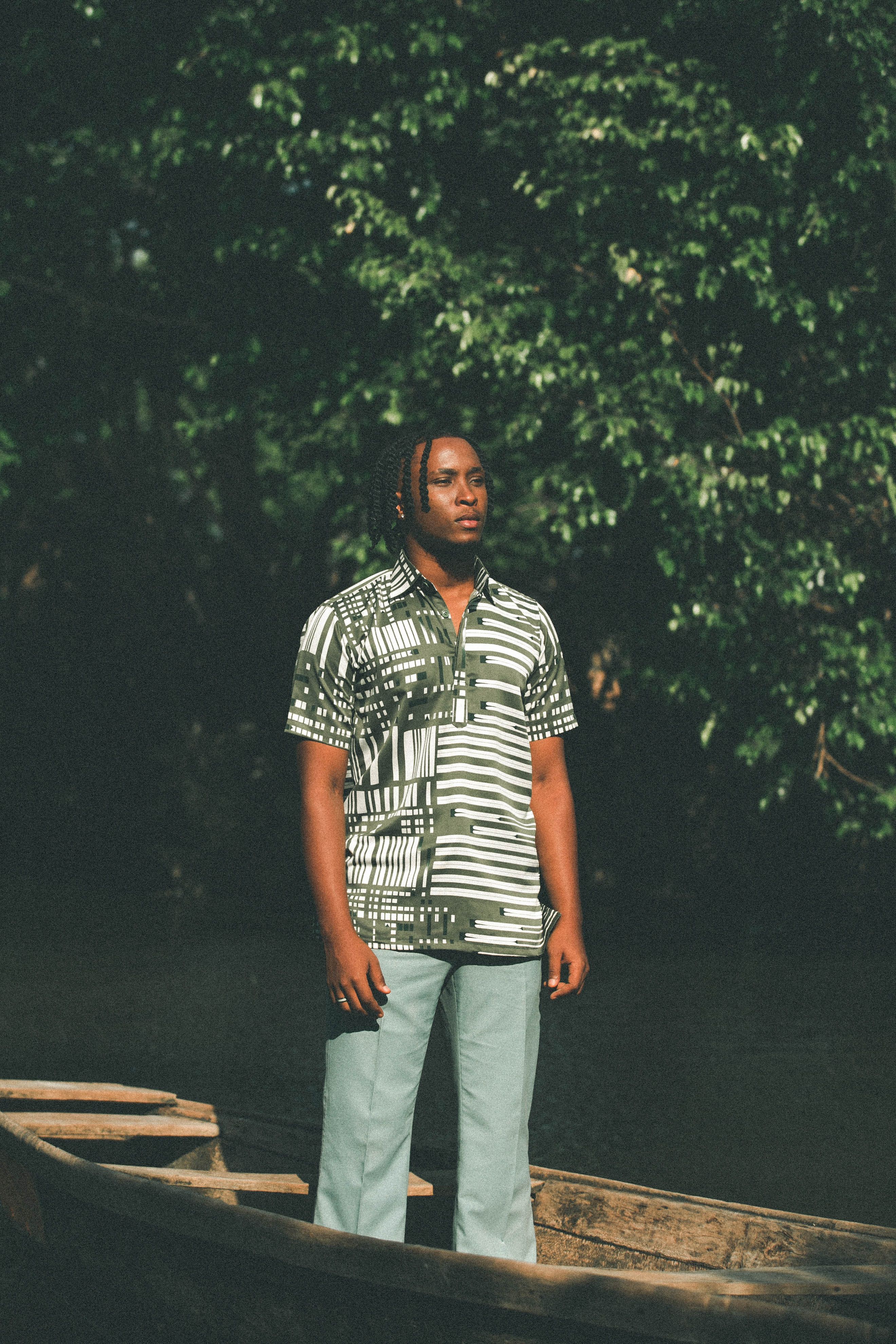Man in patterned shirt stands confidently in a wooden boat surrounded by lush greenery.