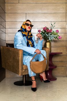 Elegant model in blue outfit with scarf and sunglasses, sitting indoors with floral decor.
