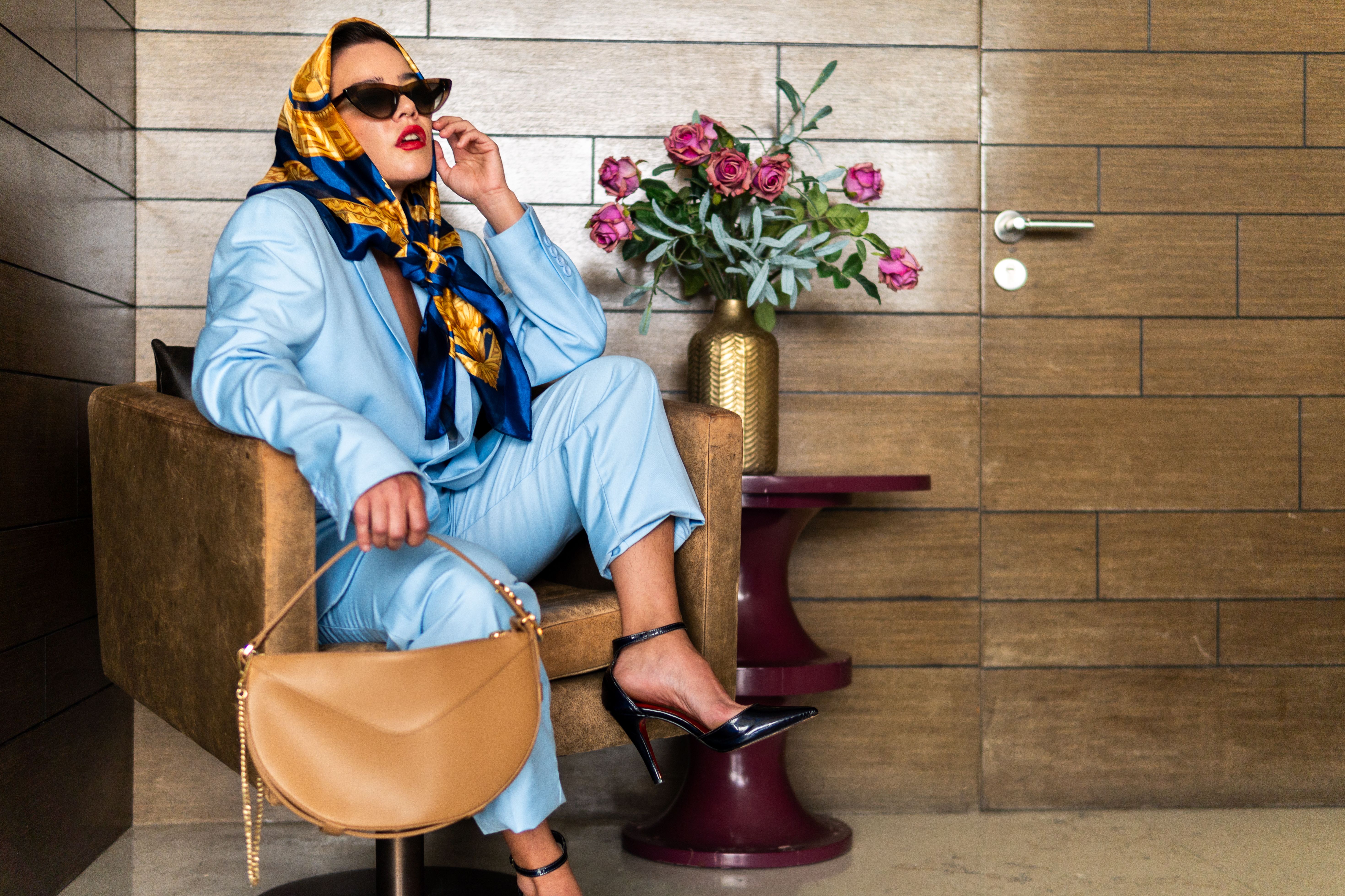 Stylish woman in a blue outfit and scarf, with sunglasses and high heels, posing indoors.