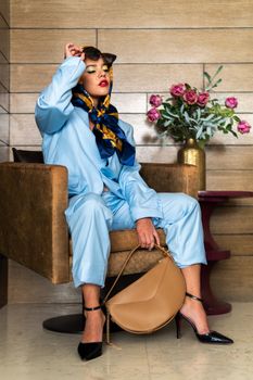 Fashionable woman in blue attire and sunglasses sitting indoors, embodying vibrant style.