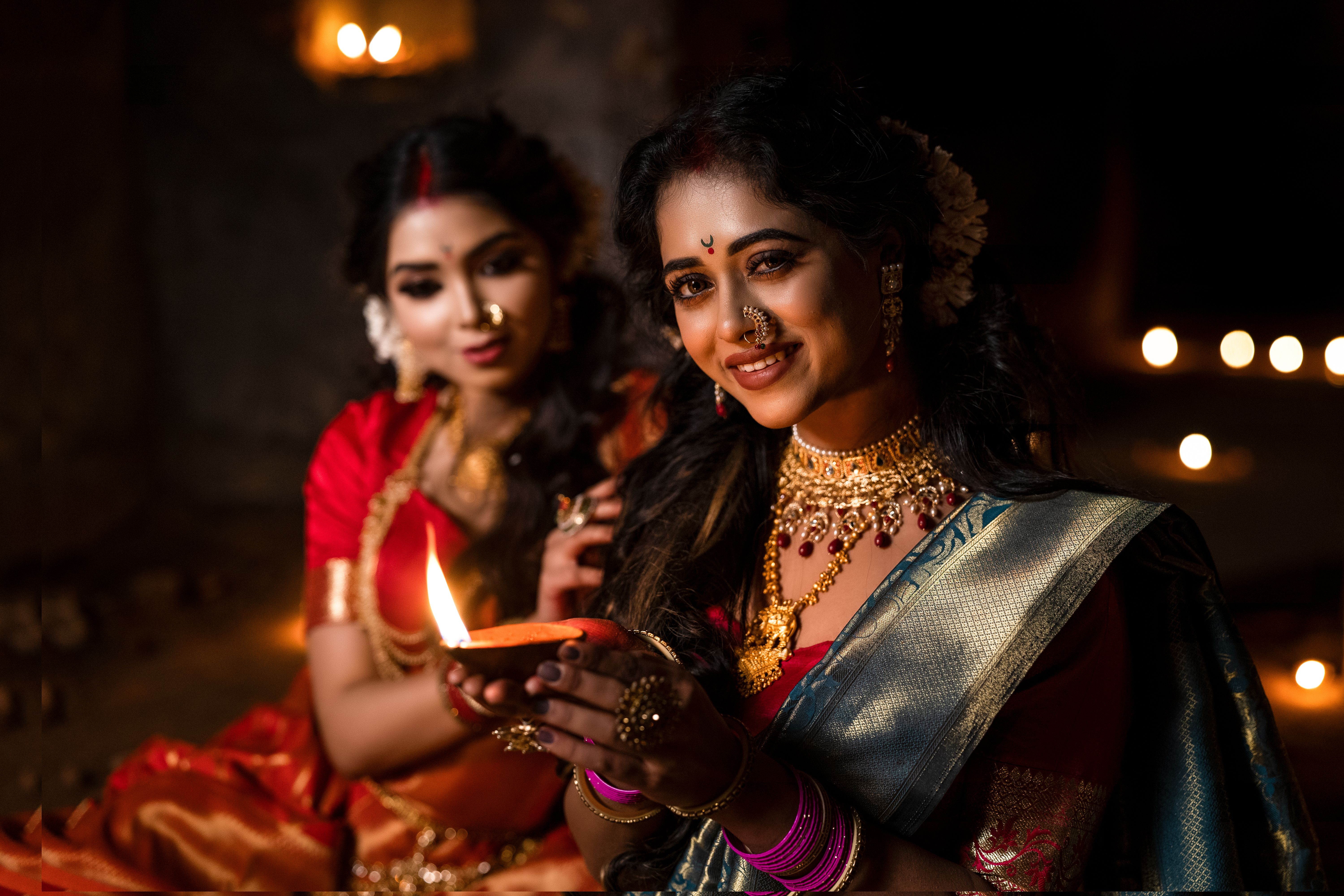 Mulheres vestidas com roupas tradicionais indianas realizando um ritual com vela em um ambiente decorado para celebração cultural ou religiosa.