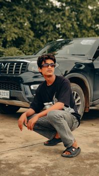 Young man squatting in front of SUV in Boraj, India, showcasing casual style and adventure.