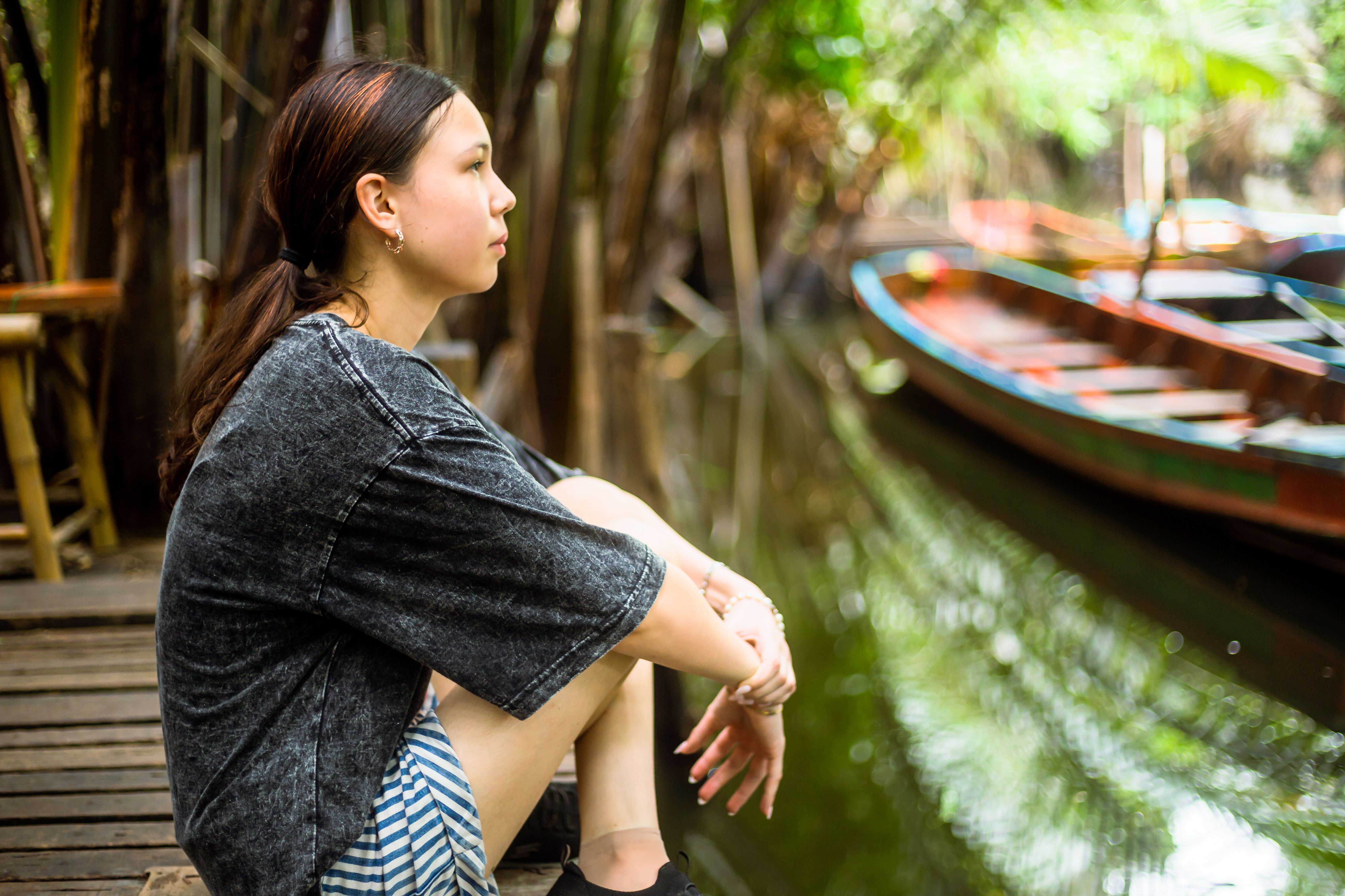 A young woman sitting on a wooden deck by a canal with colorful boats, enjoying a quiet moment.