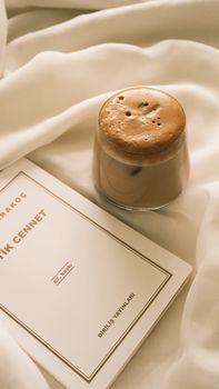 Warm cappuccino with frothy top beside a book on cream-colored fabric, exuding relaxation.