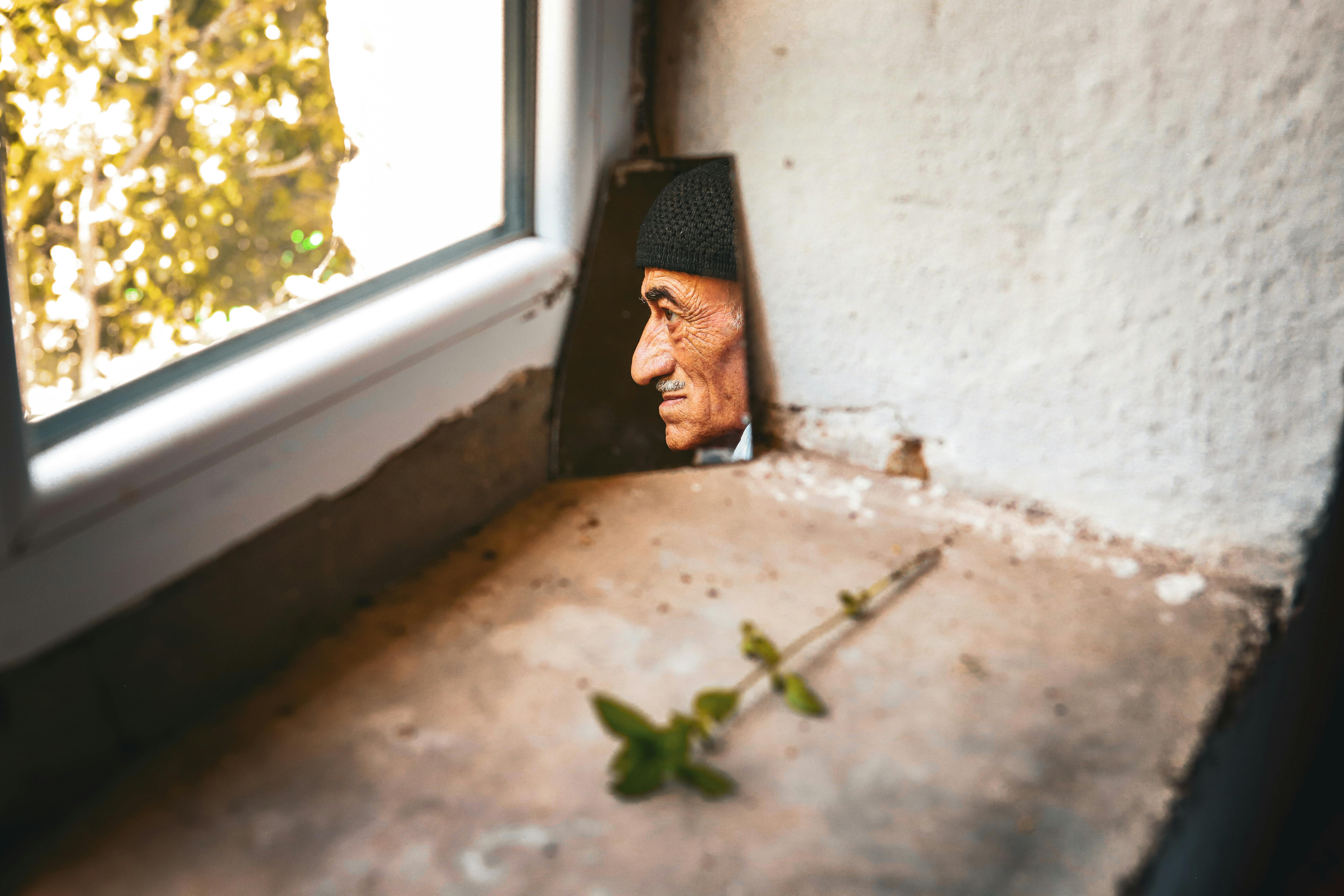 An elderly man's face is seen in reflection by a window ledge with natural light.